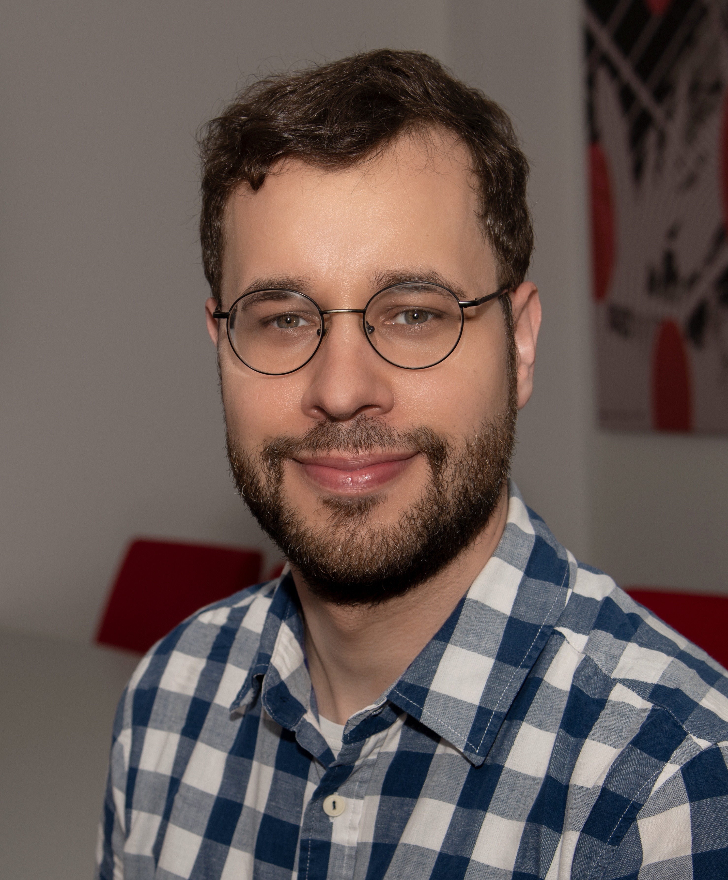 Porträt eines Mannes mit Brille und Bart, der ein blau-weiß kariertes Hemd trägt und lächelt. Im Hintergrund ist eine Wand mit einem abstrakten Kunstwerk zu sehen.